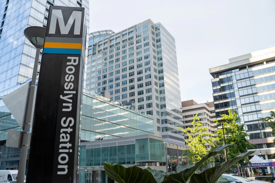 Close-up of Metro sign column at Rosslyn Station in Alexandria, Virginia with high-rose glass buildings in background.